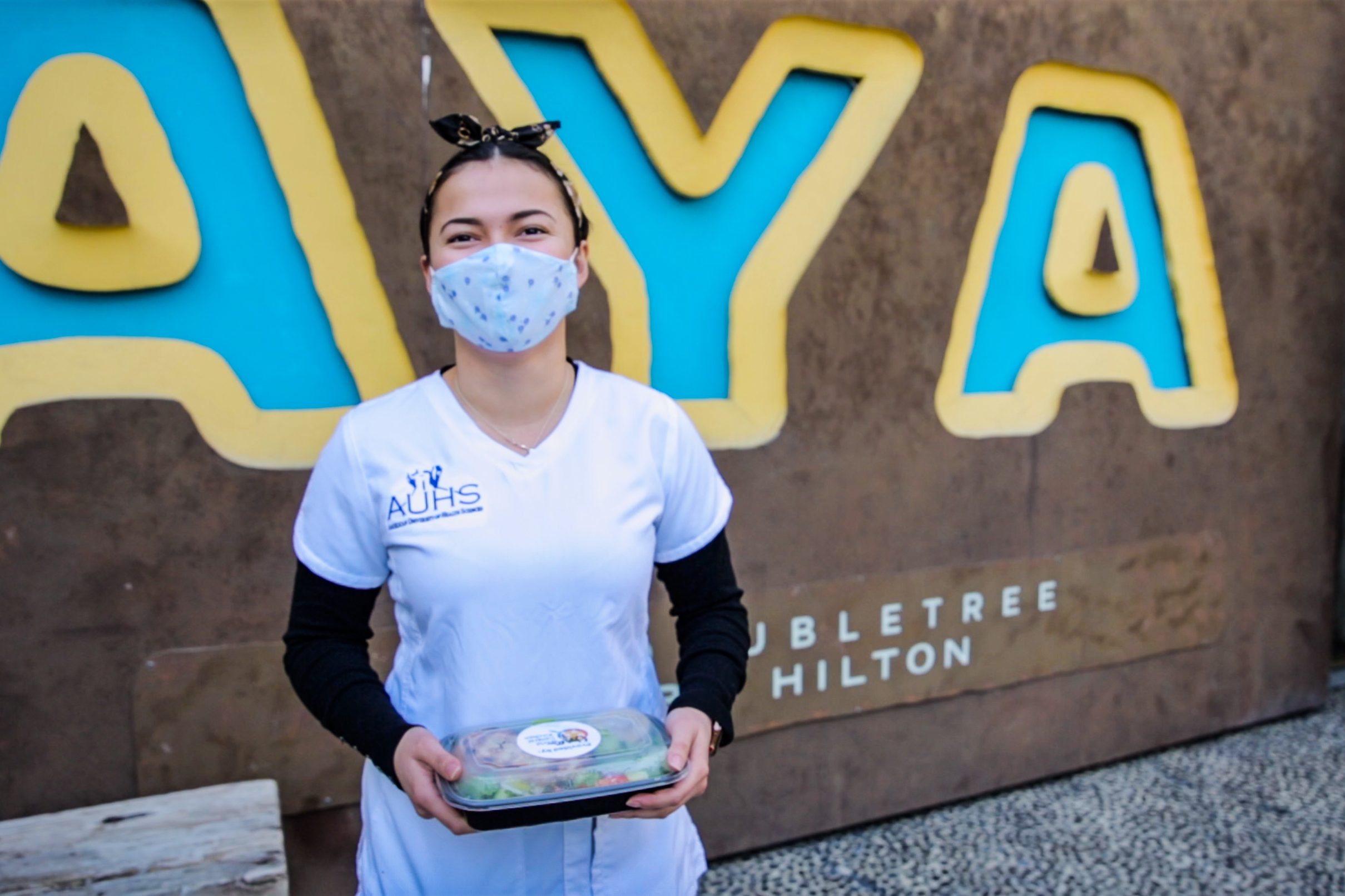 A woman wearing a face mask holds a boxed lunch in front of a wall.
